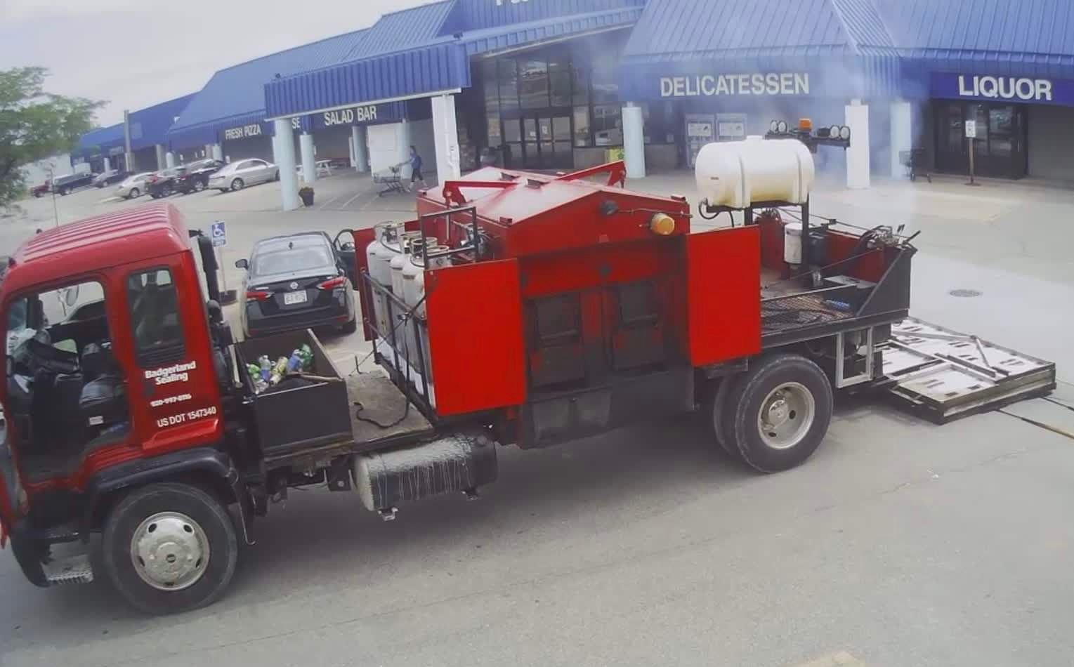 Red asphalt repair truck at a shopping center. Smoke visible.