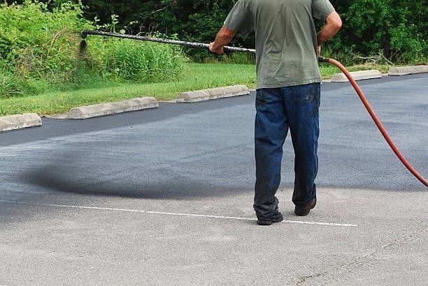 Man applying black sealant to asphalt parking lot with a long sprayer hose.
