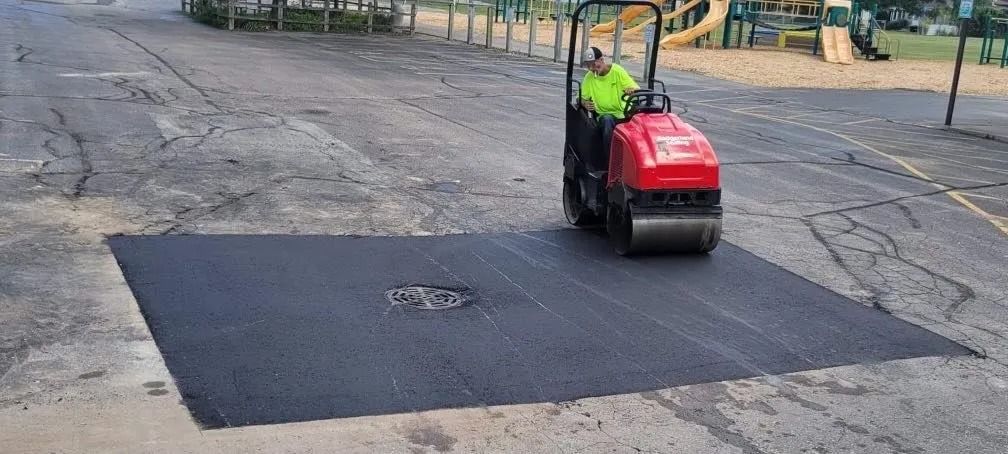 A worker operating a red asphalt roller on a freshly paved patch of road, next to a playground.