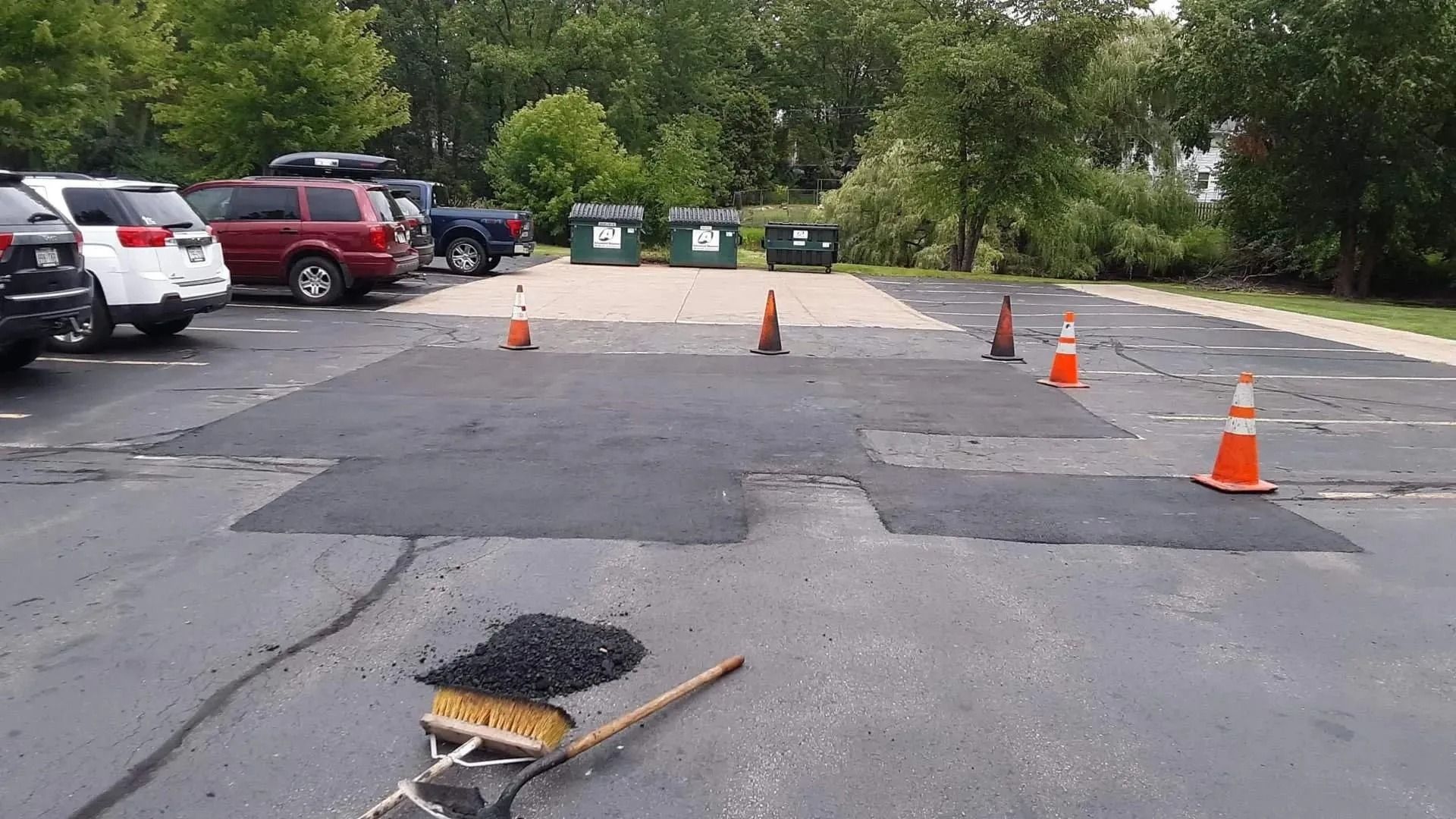 Parking lot with fresh asphalt patches, bordered by orange cones. Dumpsters and vehicles in the background.