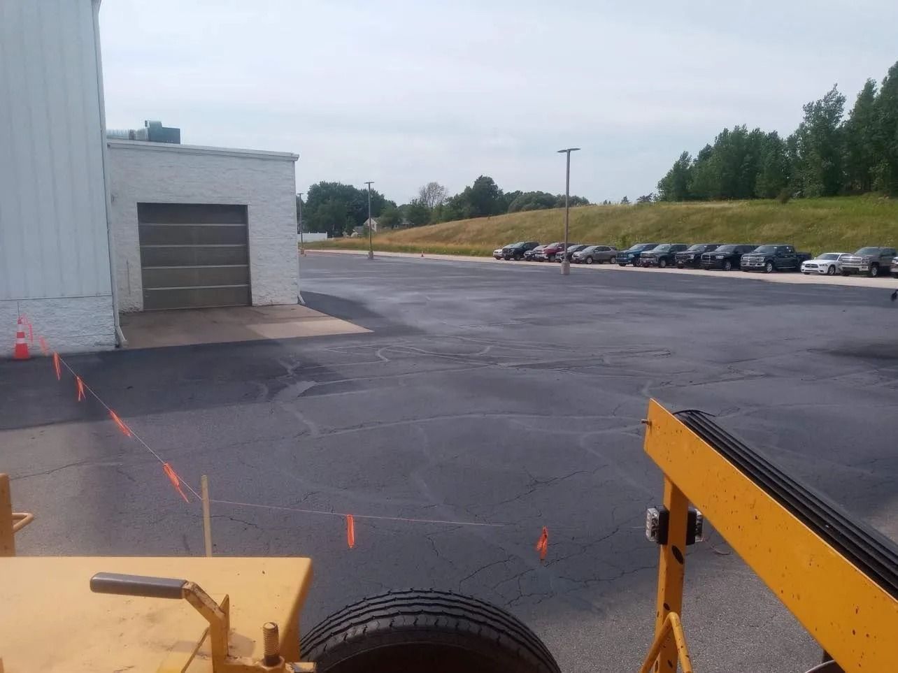 Black asphalt parking lot with parked cars, a building with a closed garage door, and orange safety cones.