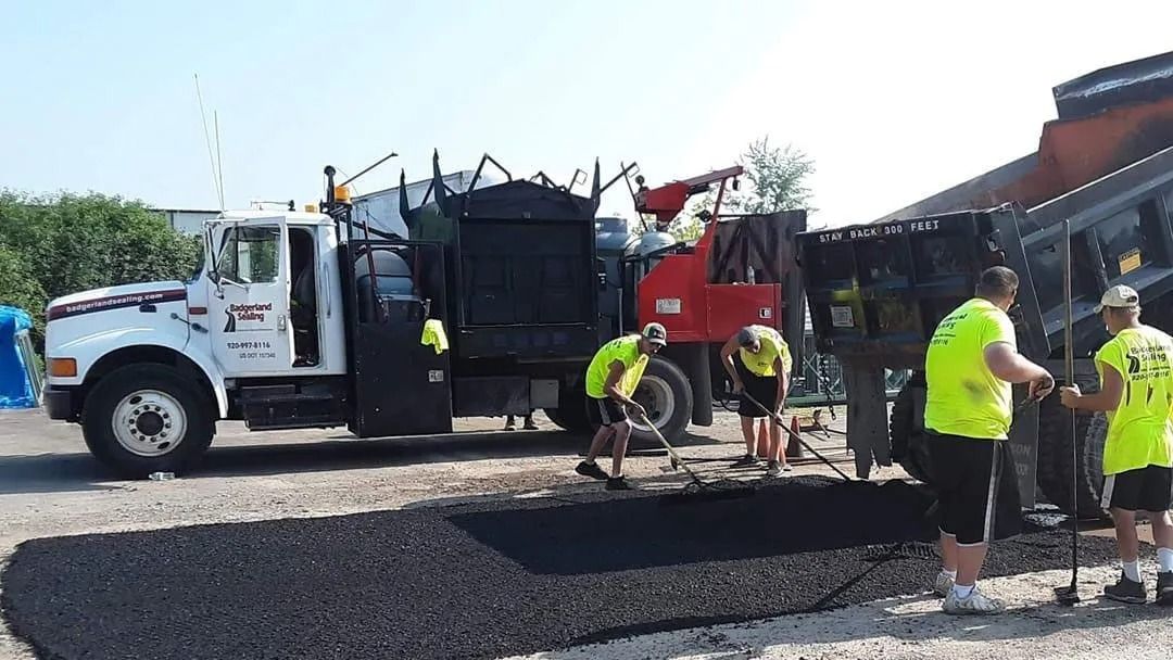 Road construction: Crew laying asphalt from truck. Men in safety vests work outdoors, using rakes and shovels.