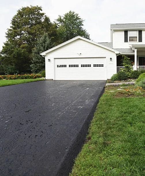 Black asphalt driveway leading to a white garage next to a lawn and two-story white house with dark shutters.