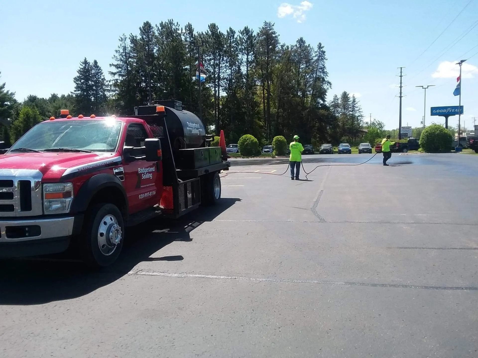 Red truck applying sealant to parking lot; two workers in neon green spray asphalt. Sunny day.