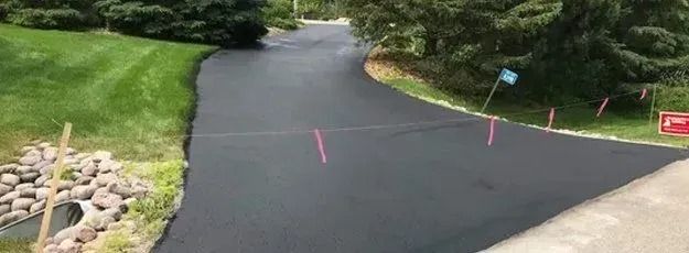 Asphalt driveway with pink stakes and string, bordered by grass and trees.