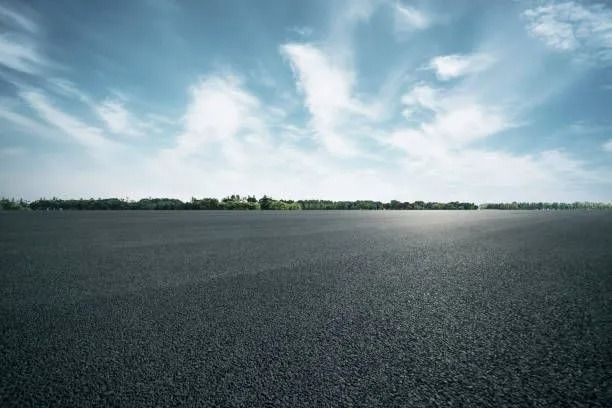 Asphalt road under a blue sky with scattered clouds, trees in the distance.