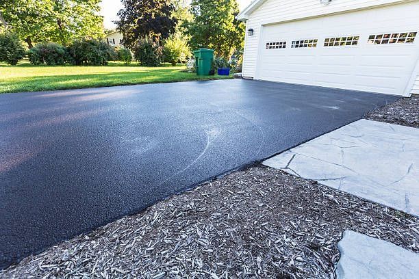 Freshly sealed black asphalt driveway next to a white garage, green lawn, and concrete pathway.