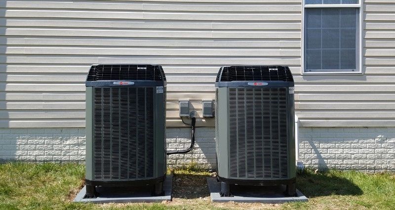 Two outdoor air conditioning units beside a house wall on a grassy lawn