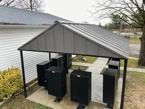 A gray metal-roofed shelter covering several black standing mailboxes on a concrete pad next to a white house.