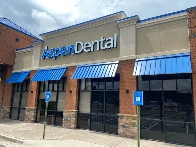 The storefront of an Aspen Dental office with beige walls, brick accents, and blue awnings under a cloudy sky.