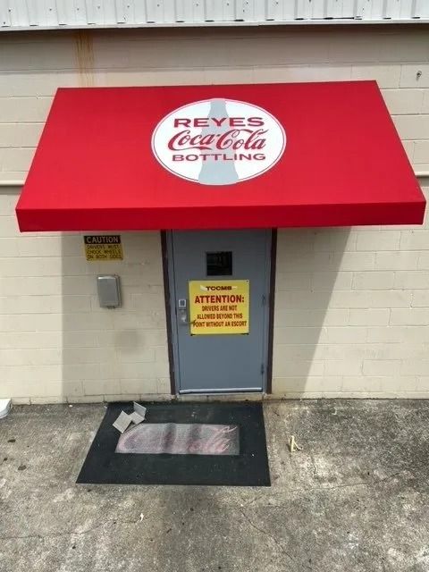 A Reyes Coca-Cola Bottling entrance with a red awning, a grey door featuring an attention sign, and a black floor mat.