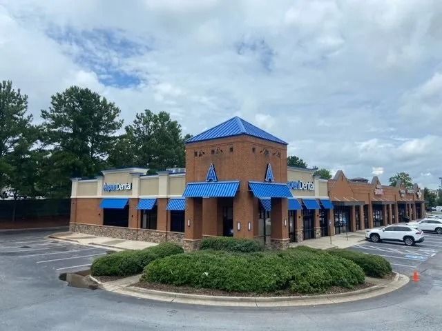 A corner view of a brown brick retail strip mall with a blue metal roof and awnings under a cloudy sky.