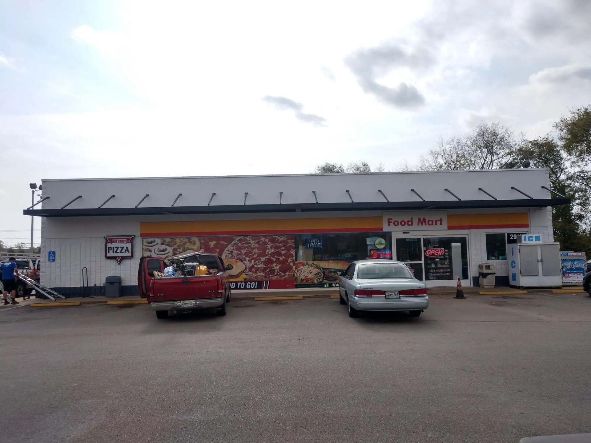 A single-story Shell gas station and food mart building under a cloudy sky, with a red truck and silver sedan parked in front.