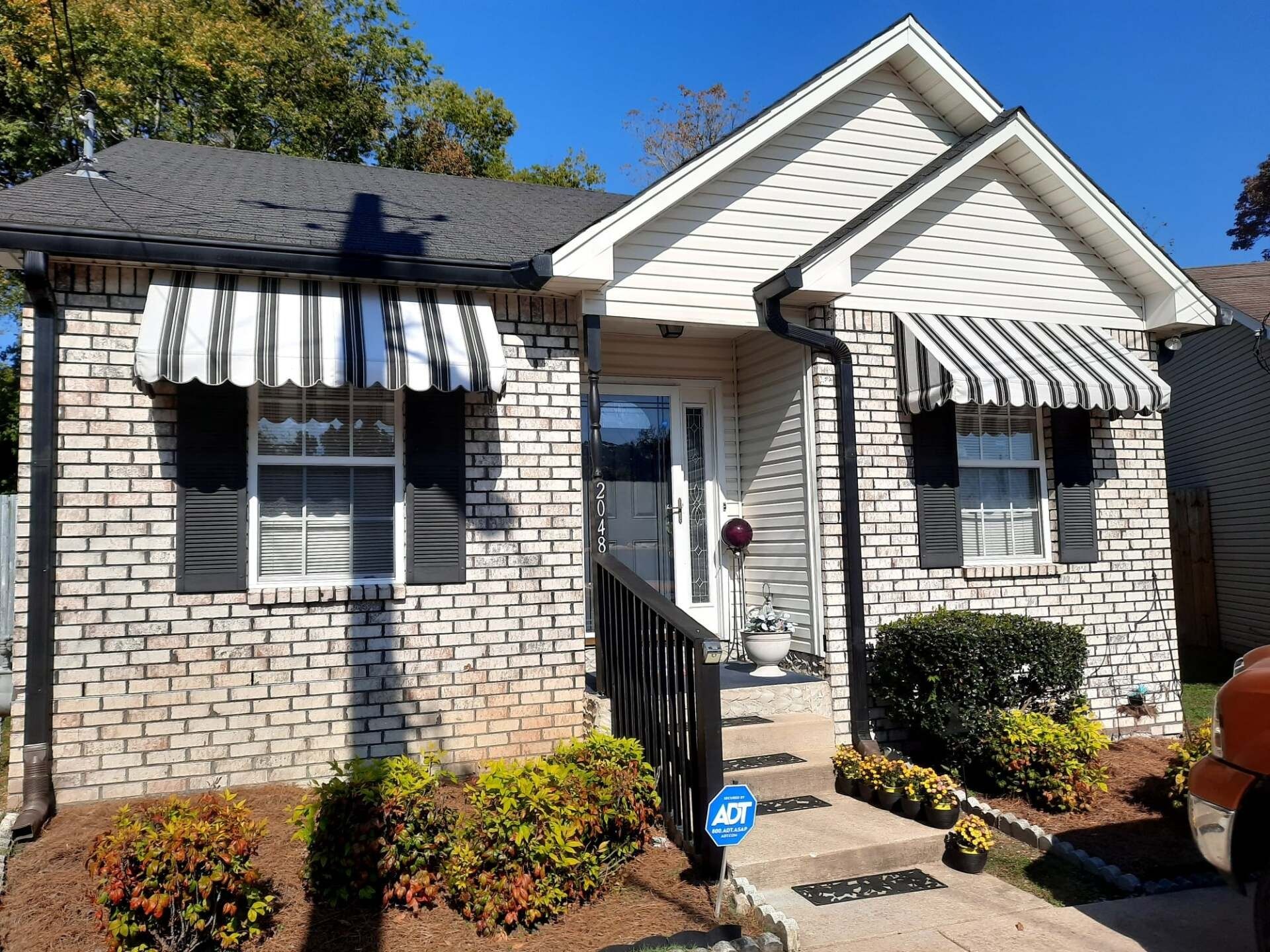 A one-story brick house with a gable roof, black-and-white striped awnings over the windows, and small landscaping bushes.