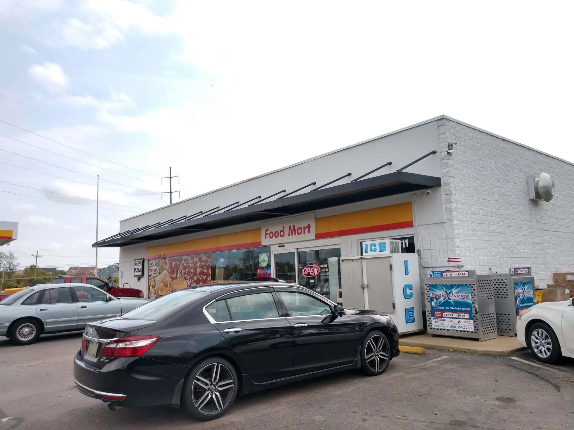 A Food Mart convenience store at a Shell gas station under a cloudy sky, with cars parked in the lot.