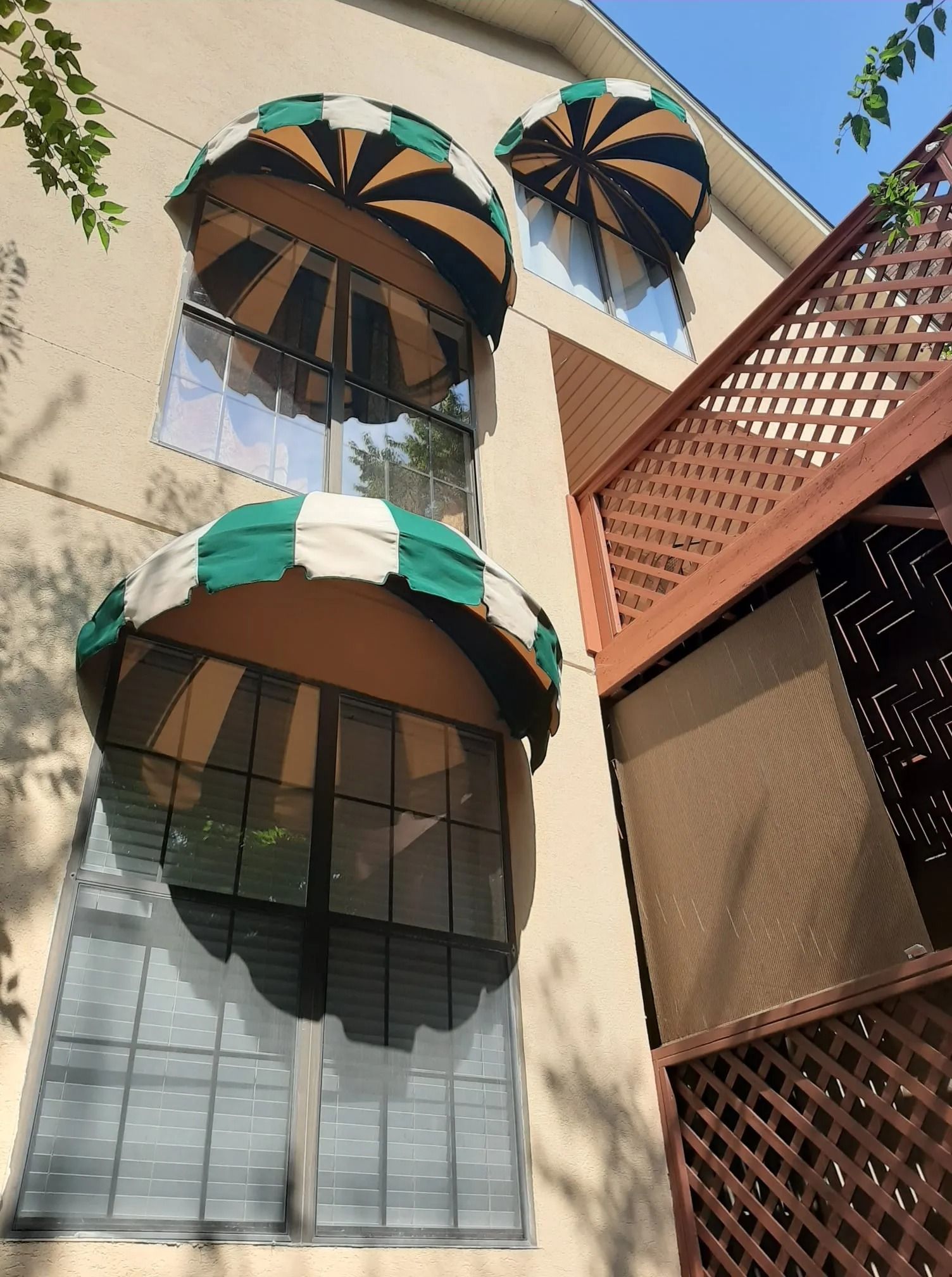 A light-colored building exterior with three green and tan striped dome-shaped window awnings and a wooden lattice deck.