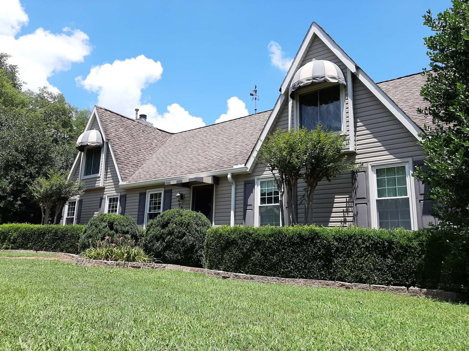 A two-story grey house with a pitched roof, front-facing gables, and dark shutters, set behind a neatly trimmed hedge.