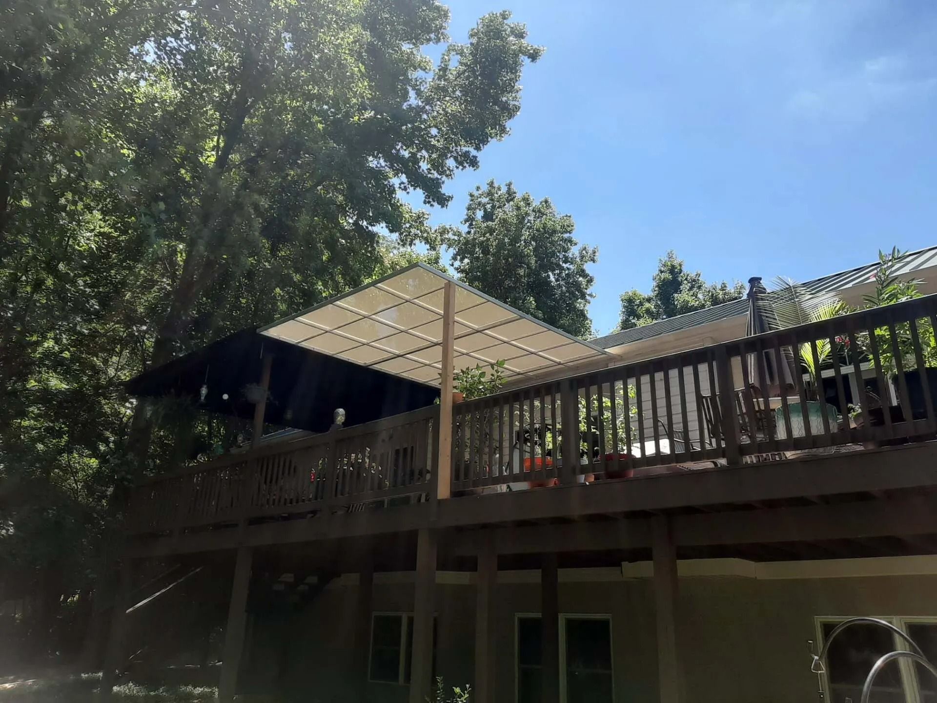 A wooden backyard deck with a light-colored, square shade canopy held up by a central post under tall, leafy trees.