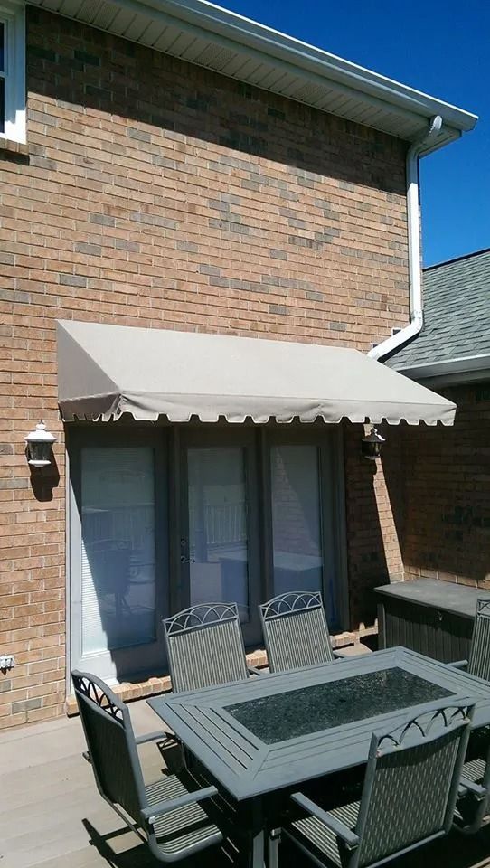 A beige fabric awning is installed above a large glass door on a brick house, shading an outdoor dining set on the patio.