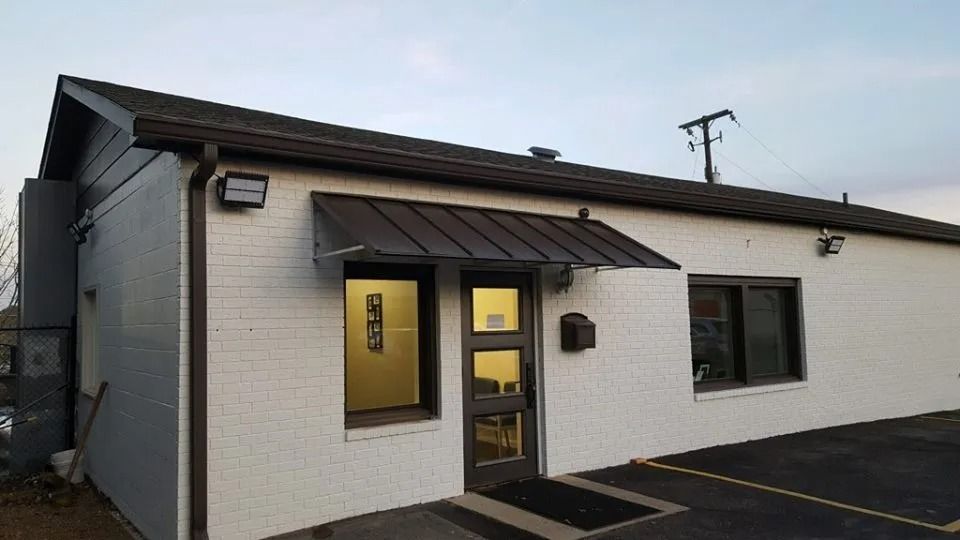 A white brick building with a dark metal awning over the entrance, featuring a glass door and two windows.