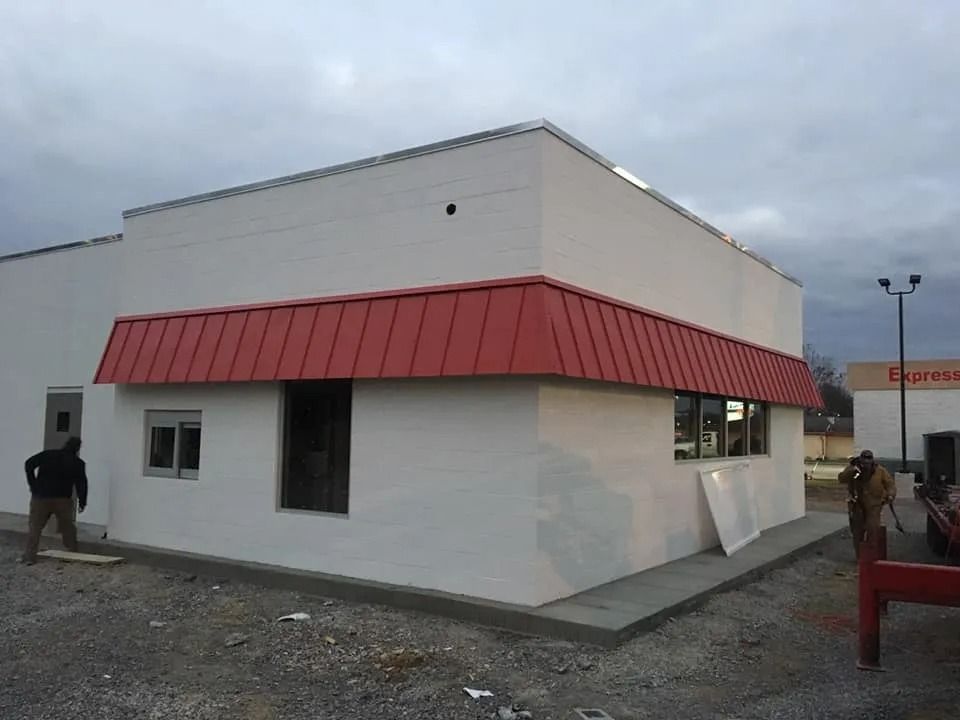 A white commercial building with a red metal awning under construction, with two workers present in a gravel lot.