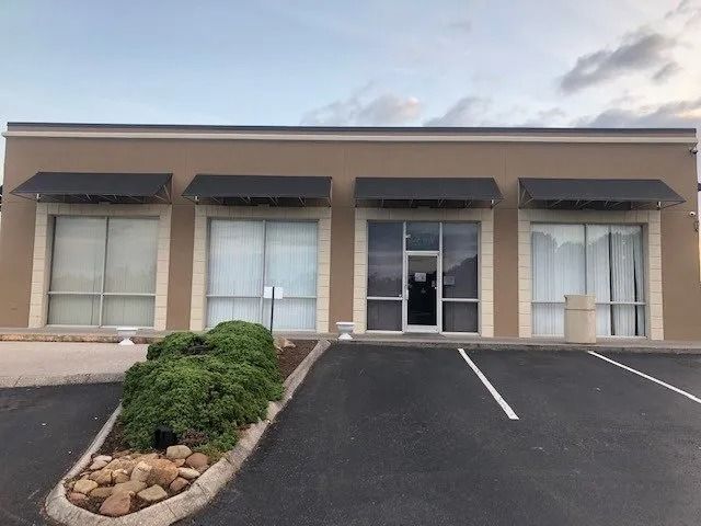 A single-story commercial building with tan walls, dark awnings, large windows, and a glass-paneled entrance.