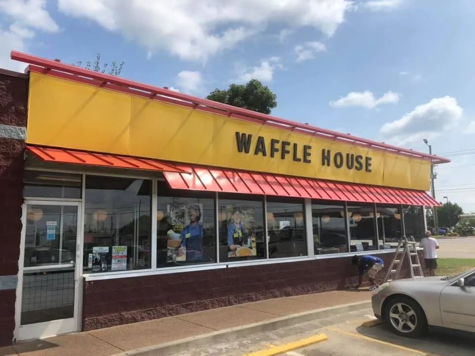 A yellow and red Waffle House restaurant exterior under a blue sky with workers near the windows and a car parked outside.