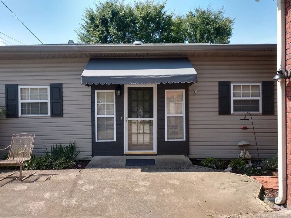 A tan house exterior with gray siding, black shutters, a dark door with side windows, and a gray fabric door awning.