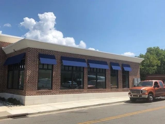 A brick commercial building with blue window awnings stands under a blue sky next to a parked orange pickup truck.
