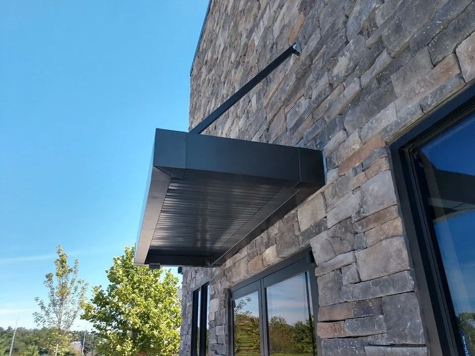 A metal awning attached to a textured stone wall above a window on a sunny day.