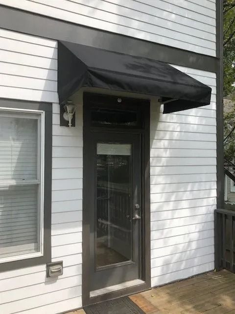 A dark-framed door with a window and a black fabric awning, set in a white horizontal-siding wall.