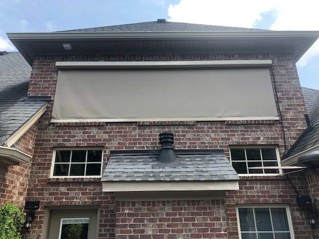 A brick house exterior featuring a large, tan retractable solar shade installed above two windows and a roof bump-out.