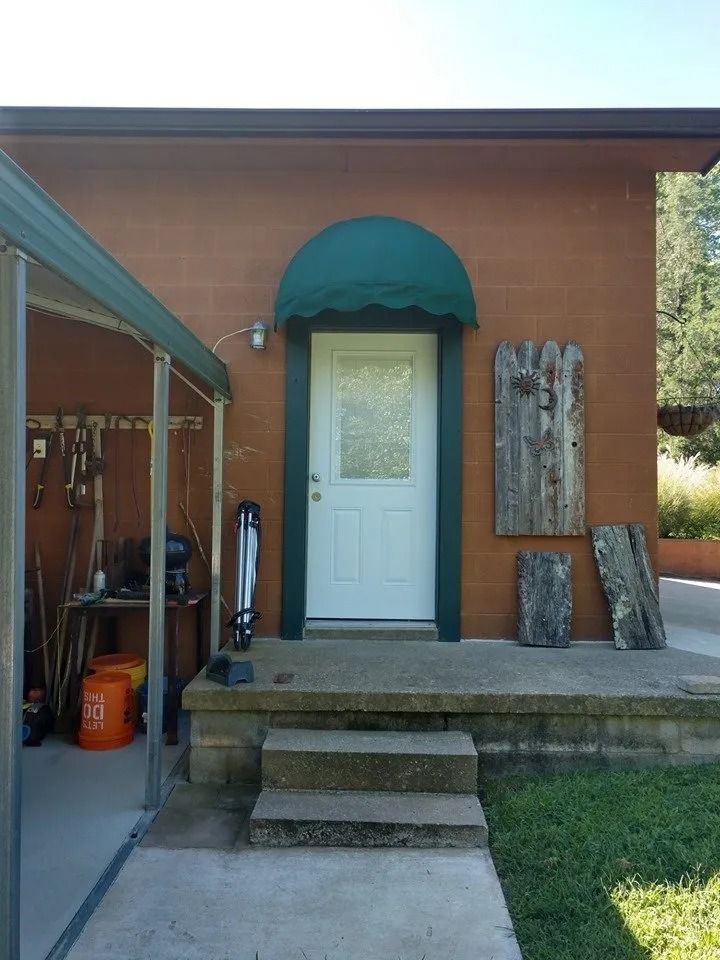 A white door with a green awning and dark trim, centered on a tan exterior wall with concrete steps and a side carport.