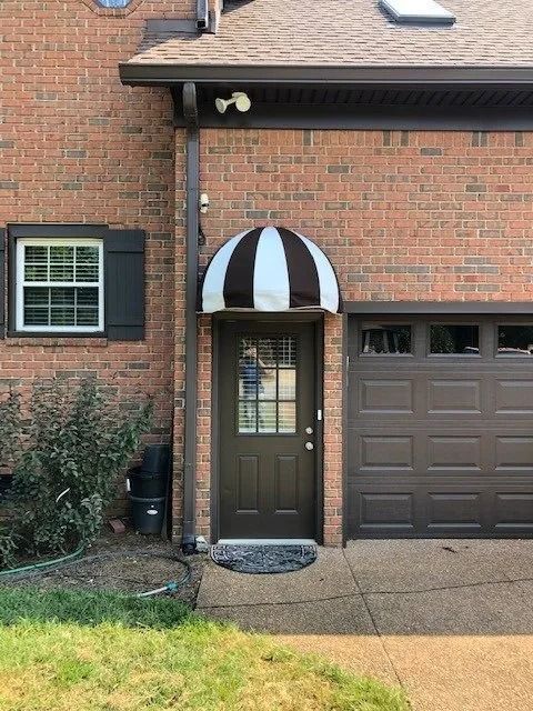 A brown brick home exterior with a dark brown door, a striped dome awning, a window, and a dark garage door.
