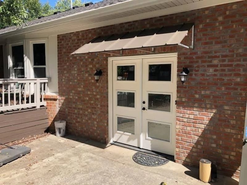Exterior brick wall featuring white double doors with three windows each, topped by a metal awning and two wall lights.