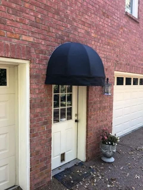 A black dome awning above a white glass-paneled door set in a brick wall next to a garage door.