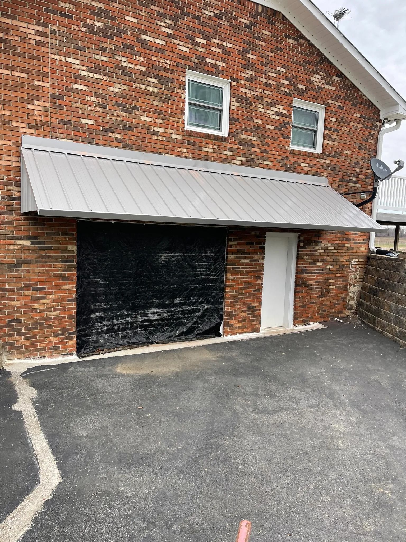 A brick building exterior with a metal awning over a large, covered black garage door and an adjacent white entry door.