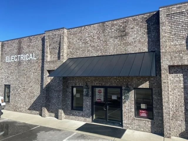 A brick building exterior with an electrical business sign, a black metal awning, and double glass entrance doors.