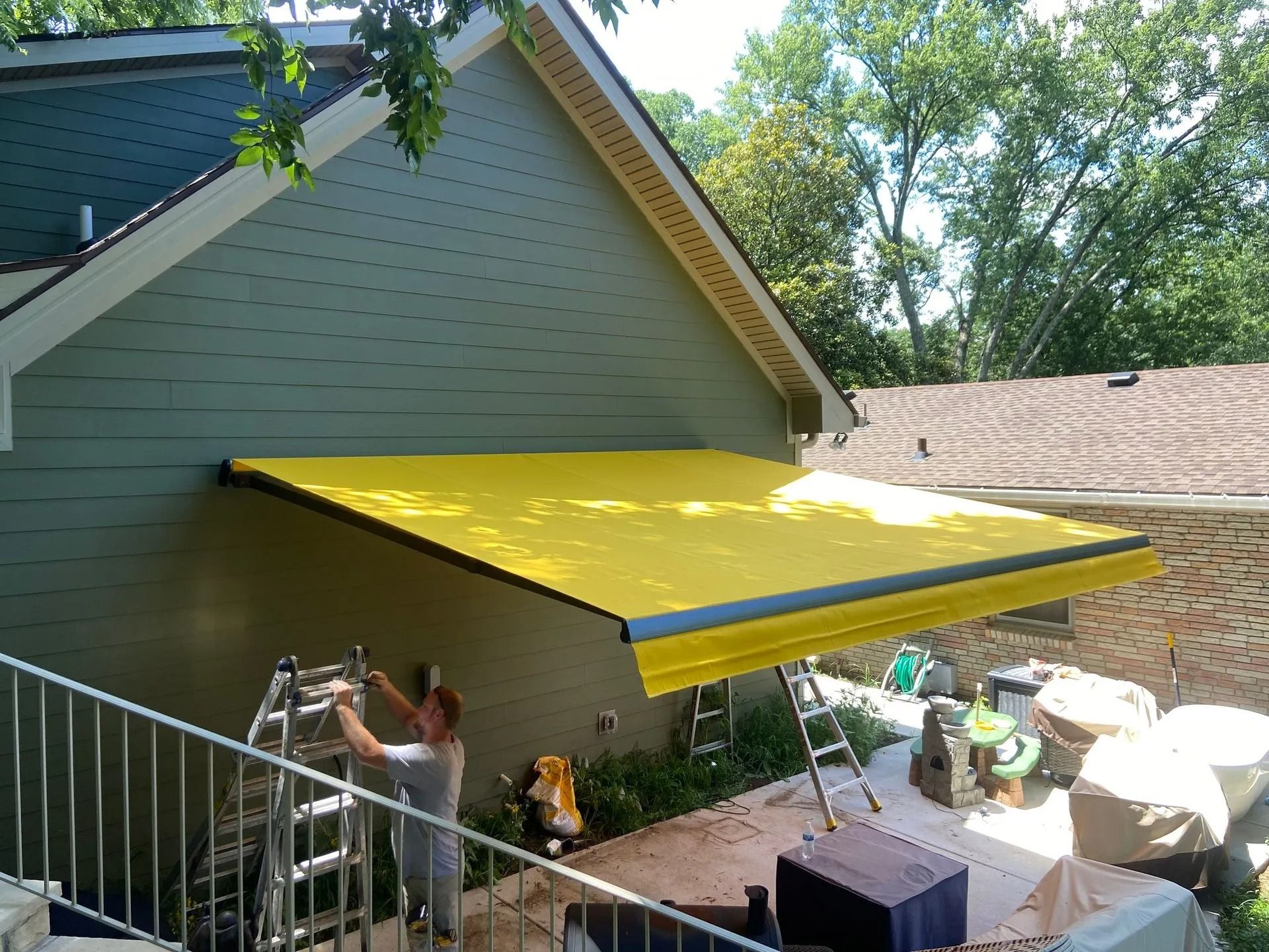A person on a ladder installs a bright yellow retractable awning on the side of a grey house.