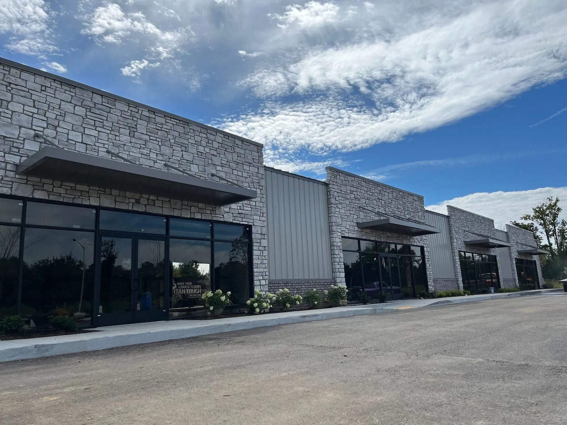 A row of modern commercial units with white stone facades, large glass storefronts, and dark awnings under a blue sky.