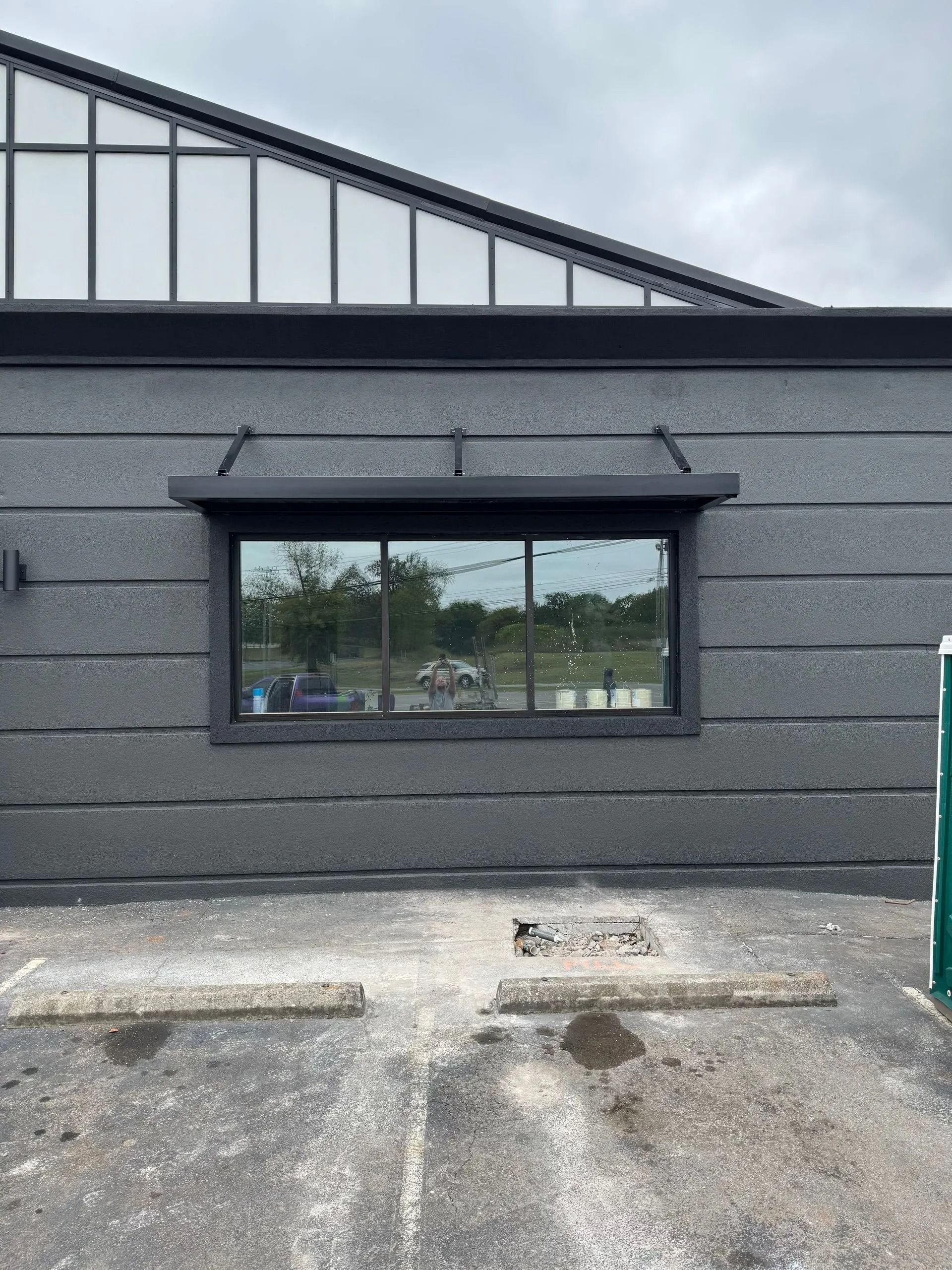 A dark grey building exterior featuring a wide horizontal window with a metal awning, viewed from a parking lot.