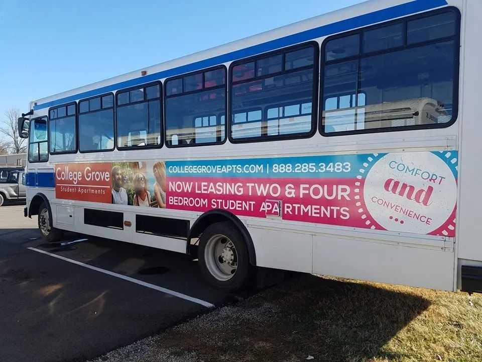 A white shuttle bus parked on asphalt featuring a promotional advertisement for College Grove student apartments.