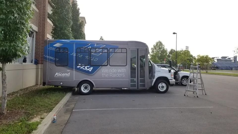 A grey and blue bus parked in a parking lot next to a building, with a stepladder nearby.