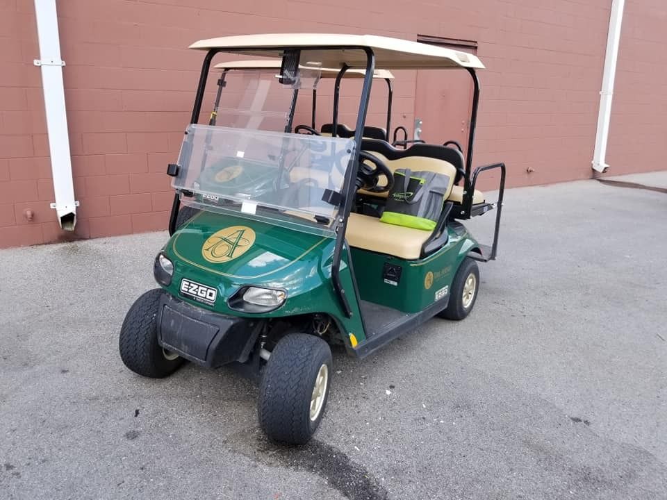 A green E-Z-GO golf cart with a beige roof and seats parked on a gray asphalt surface in front of a brown wall.