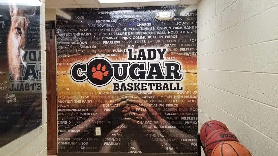 Wall graphic for Lady Cougar Basketball featuring a paw print, hands held together, and words over an orange gradient.