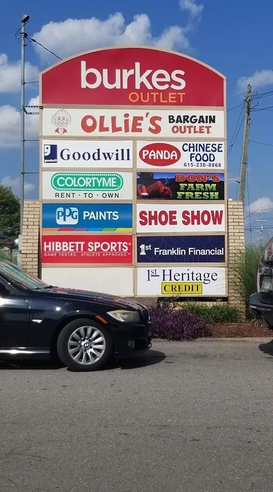A tall, multi-tenant outdoor sign for a shopping center under a blue sky, partially obscured by a parked black car.