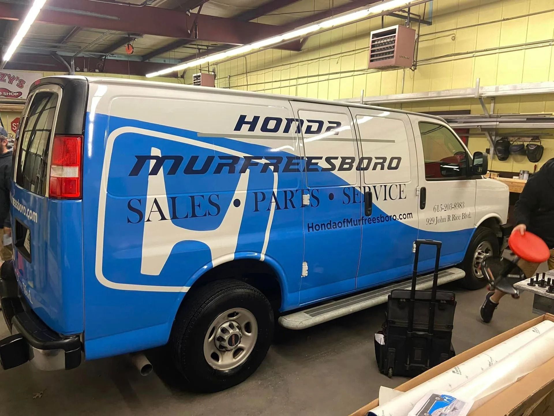 A blue and white Honda of Murfreesboro service van parked inside a workshop with a person standing nearby.