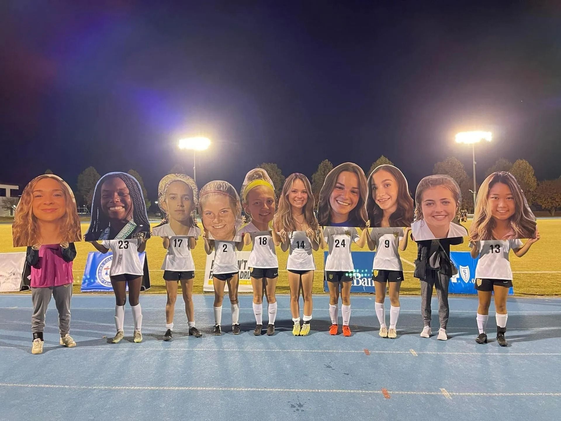 A line of ten soccer players on a blue track at night, holding up large cardboard cutouts of their own smiling faces.