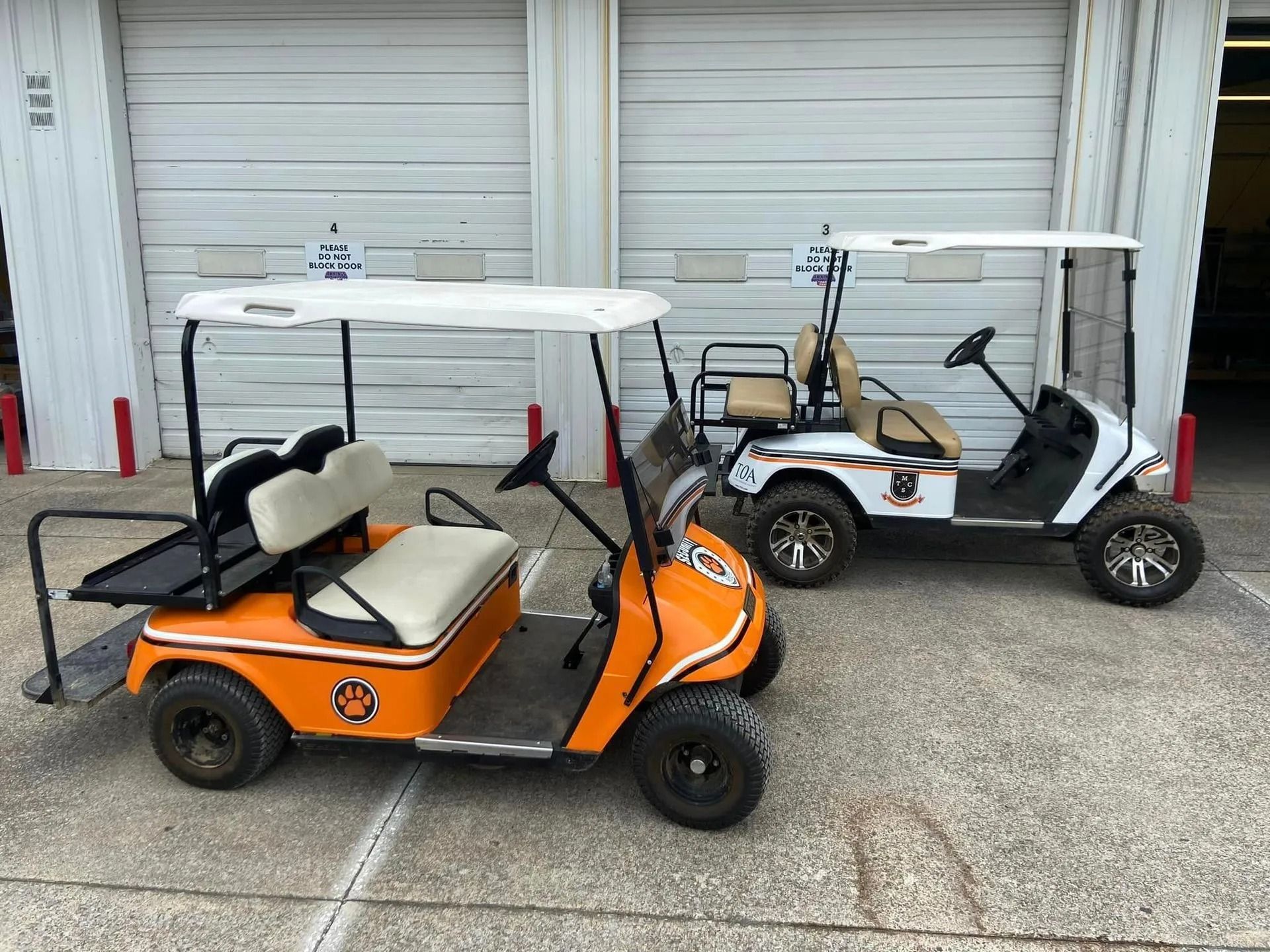 Two golf carts, one orange and one white, parked side-by-side on a concrete surface in front of metal garage doors.