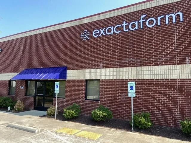The red brick building of Exactaform, featuring a blue awning over the entrance and two handicap parking signs.